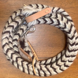 May include: A braided rope with a chevron pattern in brown and cream colors. The rope has leather accents and metal clasps. This item is likely used for equestrian activities.