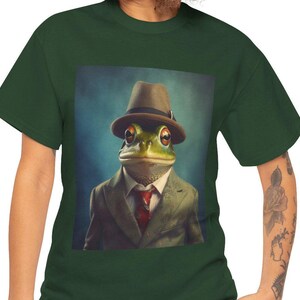 Könnte beinhalten: Grünes T-Shirt mit einer humorvollen Illustration einer Froschfigur in Anzug und Hut. Der Frosch trägt einen braunen Hut, einen grünen Anzug und eine rote Krawatte. Der Hintergrund ist blau. Das Bild ist originell.