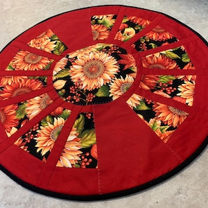Puede incluir: Una falda de mesa redonda roja con ribete negro. La falda está decorada con un patrón de girasol en tonos naranja, amarillo y verde.