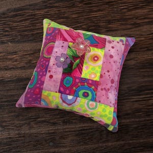 以下が含まれることがあります： 鮮やかな花柄のデザインが施された、正方形のパッチワークのピンクッション。 ピンク、紫、緑色の生地に様々な模様が施されています。 2つの装飾的な花の装飾がクッションの上部に取り付けられています。