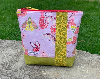 May include: A lavender zippered pouch with a yellow and green lace trim. The pouch is decorated with a butterfly and moth print. The pouch has a label that reads "Quarter Acre Arts".