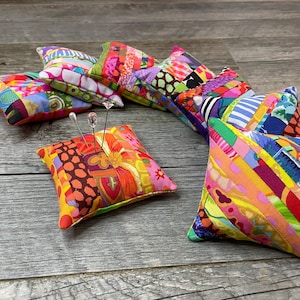 以下が含まれることがあります： 木目調の表面に、カラフルなパッチワークの針刺しが半円形に並んでいます。一部の針刺しには針が刺さっています。