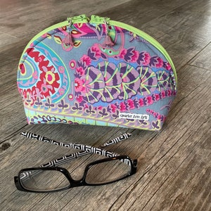 May include: A half-circle shaped zippered pouch with a paisley print in shades of pink, purple, blue, green, and yellow. The pouch has a lime green zipper and a small label that reads "Quarter Acre Arts".