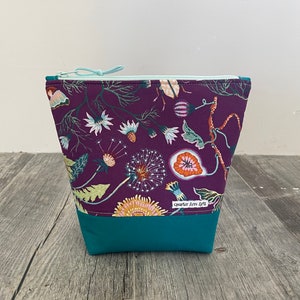 Può includere: Una pochette con cerniera viola con fondo turchese. La pochette è decorata con un motivo floreale con dente di leone, foglie e altri fiori. La pochette ha un'etichetta che recita "Quarter Acre Arts".