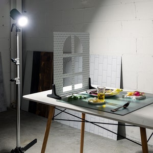 May include: A white table with a white surface and wooden legs. A green placemat with a glass of liquid, a plate with food, and a fork on it. A white backdrop with a grid pattern is behind the table.