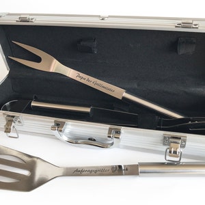 Può includere: Una valigetta di trasporto in alluminio argentato con interno nero contiene un set di tre utensili per grigliare. Gli utensili includono una spatola, delle pinze e una forchetta. La spatola e la forchetta hanno un testo inciso che recita "Auftragsgriller Frank" e "Papa der Grillmeister".