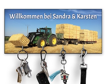 Op de afbeelding: Een sleutelbord met een foto van een groene tractor die hooibalen vervoert. De tekst "Willkommen bei Sandra & Karsten" staat op het bord gedrukt. Er zijn vier haken waaraan sleutels en een riem hangen.