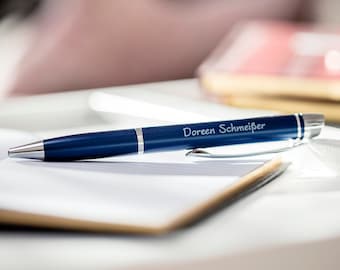 May include: A blue pen with silver accents and the name "Doreen Schmeißer" engraved on the side. The pen is lying on a white notebook.
