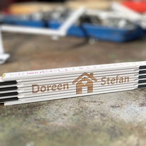 Könnte beinhalten: Ein weißes, faltbares Lineal aus Holz mit schwarzen Metallverzierungen und den aufgedruckten Namen "Doreen" und "Stefan". Das Lineal hat ein Haussymbol mit einem Herzen in der Mitte.