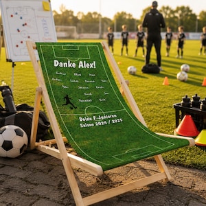 Könnte beinhalten: Ein hölzerner Klapp-Strandstuhl mit einem grünen Stoffbezug, der wie ein Fußballfeld gestaltet ist. Der Bezug trägt den Text "Danke Alex!" und Spielernamen. Der Stuhl steht auf einem Rasenfeld mit Fußbällen und Hütchen.