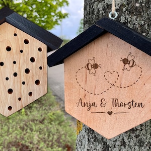 Könnte beinhalten: Ein Holz-Insektenhotel mit schwarzem Dach und einer personalisierten Inschrift "Anja & Thorsten". Zwei Bienen sind auf der Vorderseite des Hauses gezeichnet, die in Herzform fliegen.