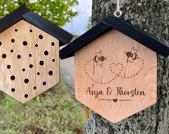 Puede incluir: Una casa de madera para abejas con un techo negro y una inscripción personalizada que dice "Anja & Thorsten". Dos abejas están dibujadas en la parte delantera de la casa, volando en forma de corazón.