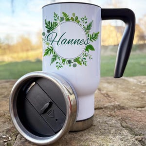 Op de afbeelding: Witte reistumbler met een zwarte deksel en handvat. De tumbler heeft een groen bladerenontwerp met een cirkel in het midden waarop "Hannes" staat.