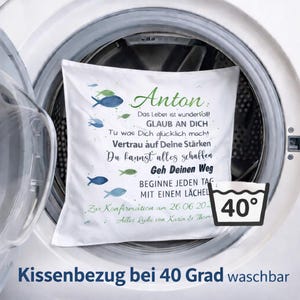 May include: A white square pillowcase featuring blue and green fish illustrations and German text including the name Anton, inspirational quotes, and a confirmation date. A black and white icon indicates the fabric is machine washable at 40 degrees.