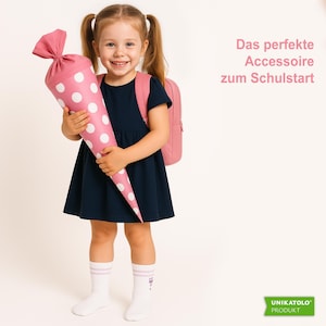 May include: A young girl smiles, holding a pink and white polka-dot school cone. She wears a navy blue dress, pink backpack, and white socks. The text on the right says, "The perfect accessory for the start of school."