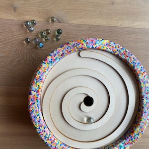 Può includere: Una pista di biglie in legno con un bordo colorato e macchiato. La pista di biglie ha la forma di una spirale e ha un piccolo foro al centro. Una biglia trasparente rotola lungo la spirale.