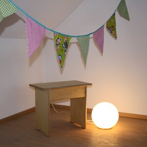 Può includere: Uno sgabello di legno con una finitura marrone chiaro si trova su un pavimento di legno. Una lampada a globo bianca è sul pavimento accanto allo sgabello. Una ghirlanda colorata con triangoli di tessuto verde, rosa e bianco è appesa al soffitto.