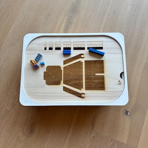 Könnte beinhalten: Ein Holztablett mit weißem Rand, mit eingravierten Umrissen einer Person und Zugwaggons. Mehrere blaue und rote Spielzeugbausteine ​​liegen auf dem Tablett. Das Tablett steht auf einer hellbraunen Holzoberfläche.