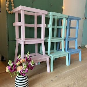 Könnte beinhalten: Vier Holztreppenhocker in verschiedenen Pastellfarben: rosa, hellgrün und blau. Die Hocker sind übereinander gestapelt, wobei der rosa Hocker unten und der blaue Hocker oben ist. Die Hocker sind aus Holz und haben ein schlichtes Design.