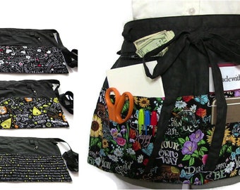 Peut inclure: Tablier utilitaire noir avec plusieurs poches et une fermeture à nouer. Le tablier a une base en toile noire avec un imprimé floral coloré et un texte inspirant. Les poches sont remplies de divers objets, notamment de l'argent liquide, un bloc-notes, des ciseaux, des stylos, un téléphone et des clés. Le texte sur le tablier est "Made with Love & Hope".