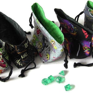 May include: Five colorful fabric dice bags with drawstring closures. The bags are lined with green fabric and feature different patterns, including a black and gold pattern, a gray and green pattern with a cartoon character, a black and purple pattern with a cartoon character, a gray and green pattern with a cartoon character, and a multi-colored pattern with a cartoon character.  Green dice are scattered in front of the bags.