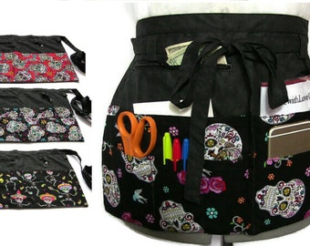 Peut inclure: Tablier utilitaire noir avec plusieurs poches et une fermeture à nouer. Le tablier présente un imprimé coloré de crânes de sucre avec des accents noirs, blancs, roses et bleus.