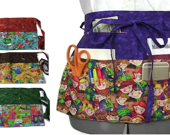 Peut inclure: Un tablier multicolore avec plusieurs poches. Le tablier a une partie supérieure violette et une partie inférieure avec un motif coloré de visages d'enfants et de globes. Le tablier a une poche zippée, une poche pour téléphone portable et un porte-clés. Le texte "Made with Love & Hopes" est imprimé sur le tablier.