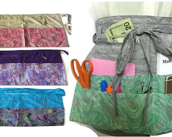 May include: Three different styles of utility aprons with multiple pockets. The aprons are made of a patterned fabric with a solid color top section. The aprons have a tie closure and are shown with various items in the pockets, including scissors, pens, a notebook, keys, and cash.