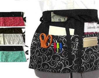 May include: Black utility apron with white swirl pattern and multiple pockets. The apron has a brown, red, and turquoise fabric trim option. The apron is shown with a notepad, scissors, pens, keys, and a phone.