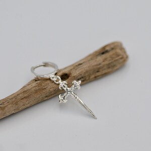 May include: Silver hoop earring with a dangling silver cross charm. The cross has a pointed tip and intricate details.