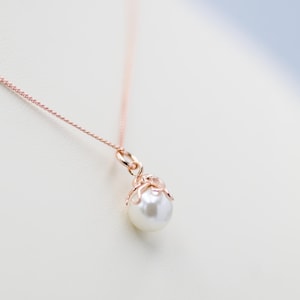 May include: A rose gold necklace with a white pearl pendant. The pendant is held in a decorative setting and hangs from a fine chain. The jewellery is shown against a soft, neutral background.