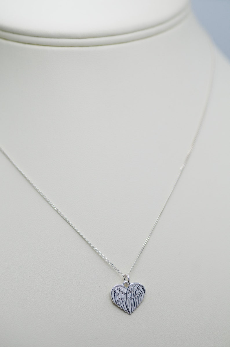 May include: A silver necklace with a heart-shaped pendant featuring a detailed angel wing design.
