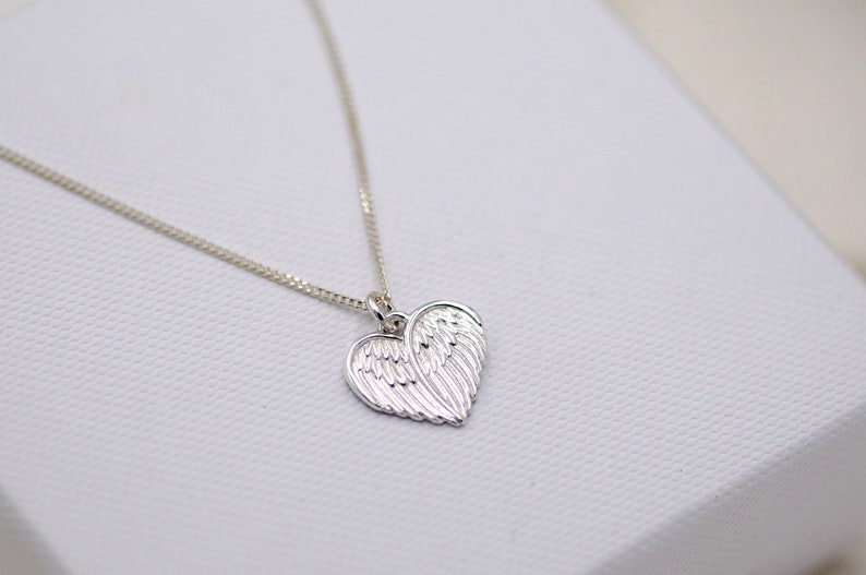 May include: A silver necklace with a heart-shaped pendant featuring a detailed design of angel wings.
