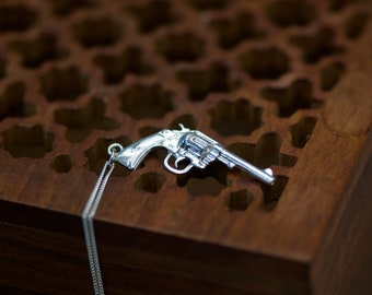 Pode incluir: Um pingente de metal prateado em forma de revólver, apresentando um cano, tambor e empunhadura detalhados. O pingente está preso a uma corrente de metal prateado fina e delicada que pende para baixo a partir do topo do berloque de arma.