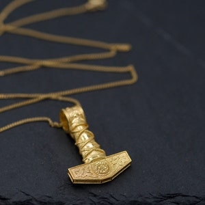 Könnte beinhalten: Eine goldene Kettenkette mit einem detaillierten Mjolnir-Anhänger. Der Anhänger ist aufwendig mit Knotenarbeiten und einem kreisförmigen Symbol auf dem Hammerkopf verziert.