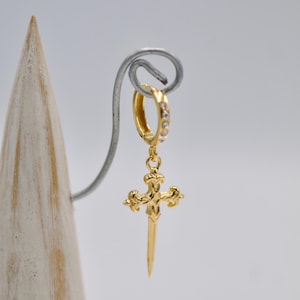 May include: Gold-toned earring with a dangling sword-shaped cross pendant. The cross has a detailed design and is accented with small clear stones. The earring is attached to a hoop with clear stones.