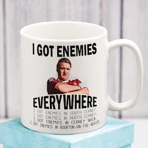 May include: White ceramic mug with the text "I GOT ENEMIES EVERYWHERE" and a picture of a man with his arms crossed. The mug also lists locations such as South Cerney and Bourton-on-the-Water.