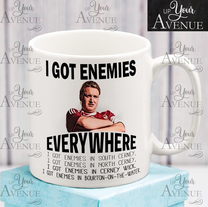 May include: White ceramic mug with the text "I GOT ENEMIES EVERYWHERE" and a picture of a man. The mug also lists locations like South Cerney and Bourton-on-the-Water. A humorous gift idea.