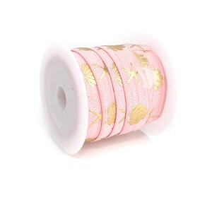 Könnte beinhalten: Eine Spule mit rosa Band mit einem sich wiederholenden Muster aus goldenen Muscheln und Seesternen. Das Band ist um eine weiße Kunststoffspule gewickelt, ideal zum Basteln oder zum Verpacken von Geschenken. Die Bandbreite beträgt ca. 1,3 cm.