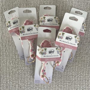 May include: A collection of Wet Brush x MacKenzie-Childs keychain detanglers. Each brush features a pink and white handle and a pink brush head. The packaging displays the text "Wet Brush x MacKenzie-Childs" and "55% less breakage for healthier looking hair."