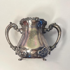 以下が含まれることがあります： 2つの装飾的なハンドルと装飾的な花の模様が付いた銀色の砂糖入れ。ボウルは溝のあるボディで、3つの小さな足の上にあります。ボウルの縁も詳細なパターンで装飾されています。