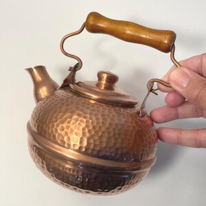 May include: A copper kettle with a wooden handle. The kettle has a hammered texture and a small spout. The handle is curved and attached to the kettle with copper wire. The lid is also copper.