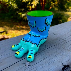 May include: A blue and teal monster-themed planter shaped like a foot with three toes. The exterior features black outlines, bright yellow toenails, and a vibrant green interior. The design includes blue and teal patches with black detailing.