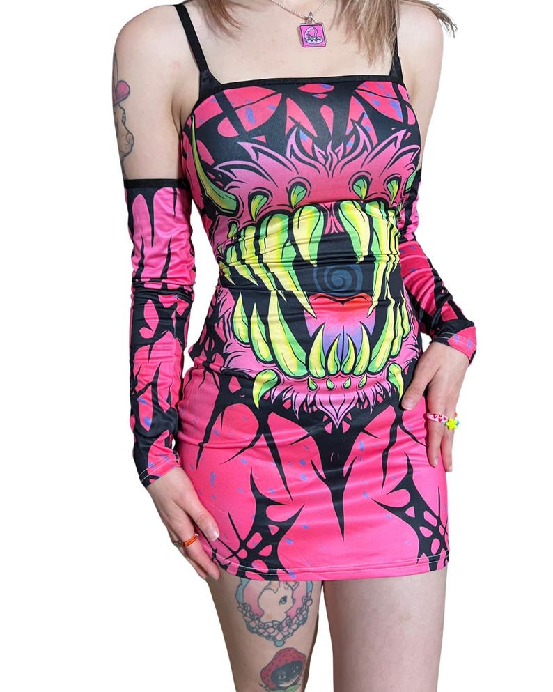 May include: A vibrant pink mini dress with a bold, monster-themed graphic. The design features a large, toothy mouth in shades of green and yellow, outlined in black. Includes matching long sleeves. The dress has a square neckline and thin straps.
