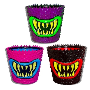 Puede incluir: Tres macetas cilíndricas con una superficie exterior texturizada y puntiaguda. Cada maceta presenta un gráfico prominente de una boca ancha con dientes amarillos afilados. La maceta superior es morada, la inferior izquierda es rosa brillante y la inferior derecha es negra.
