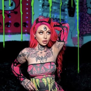 May include: A person with vibrant pink hair and elaborate tattoos, wearing a pink and black patterned tube top, matching gloves, and a mini skirt. Accessories include a horned headband and face jewelry. The background features neon green drips.
