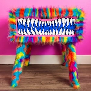 May include: A whimsical, colorful table with a pink top featuring a blue mouth design with white teeth. The table is covered in vibrant, rainbow-colored faux fur, including the legs and edges, creating a playful and unique aesthetic.