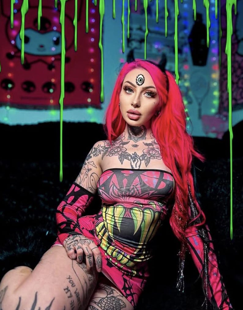 May include: A pink and black strapless dress with a matching sleeve. The dress features a colorful graphic design. The person in the image has bright pink hair and tattoos.