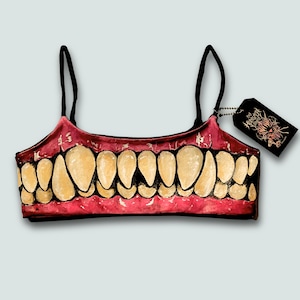 May include: A red and black crop top with a graphic design of a set of teeth. The top has black straps and a tag attached with a design and text.