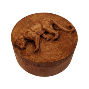 May include: A round wooden box featuring a carved relief of a large feline on the lid. The box is crafted from medium-brown wood with a visible grain pattern. The lid displays a detailed, three-dimensional depiction of a panther in a prowling pose.